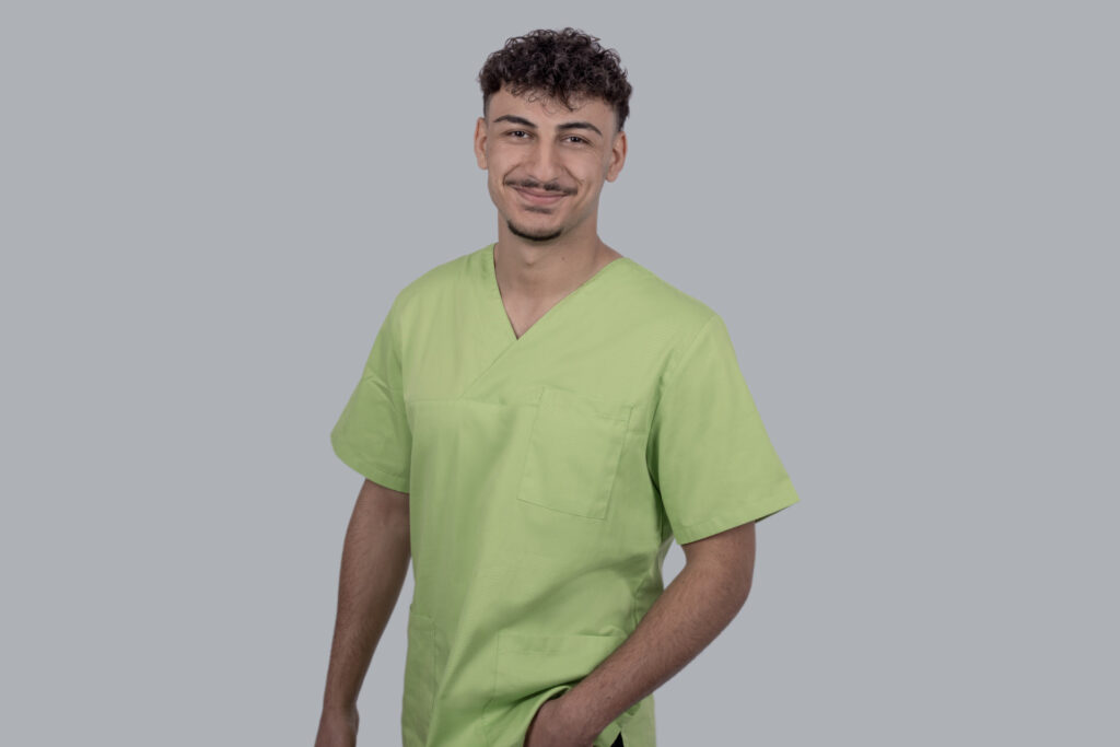 Smiling male healthcare worker in light green scrubs, torso view against gray backdrop.